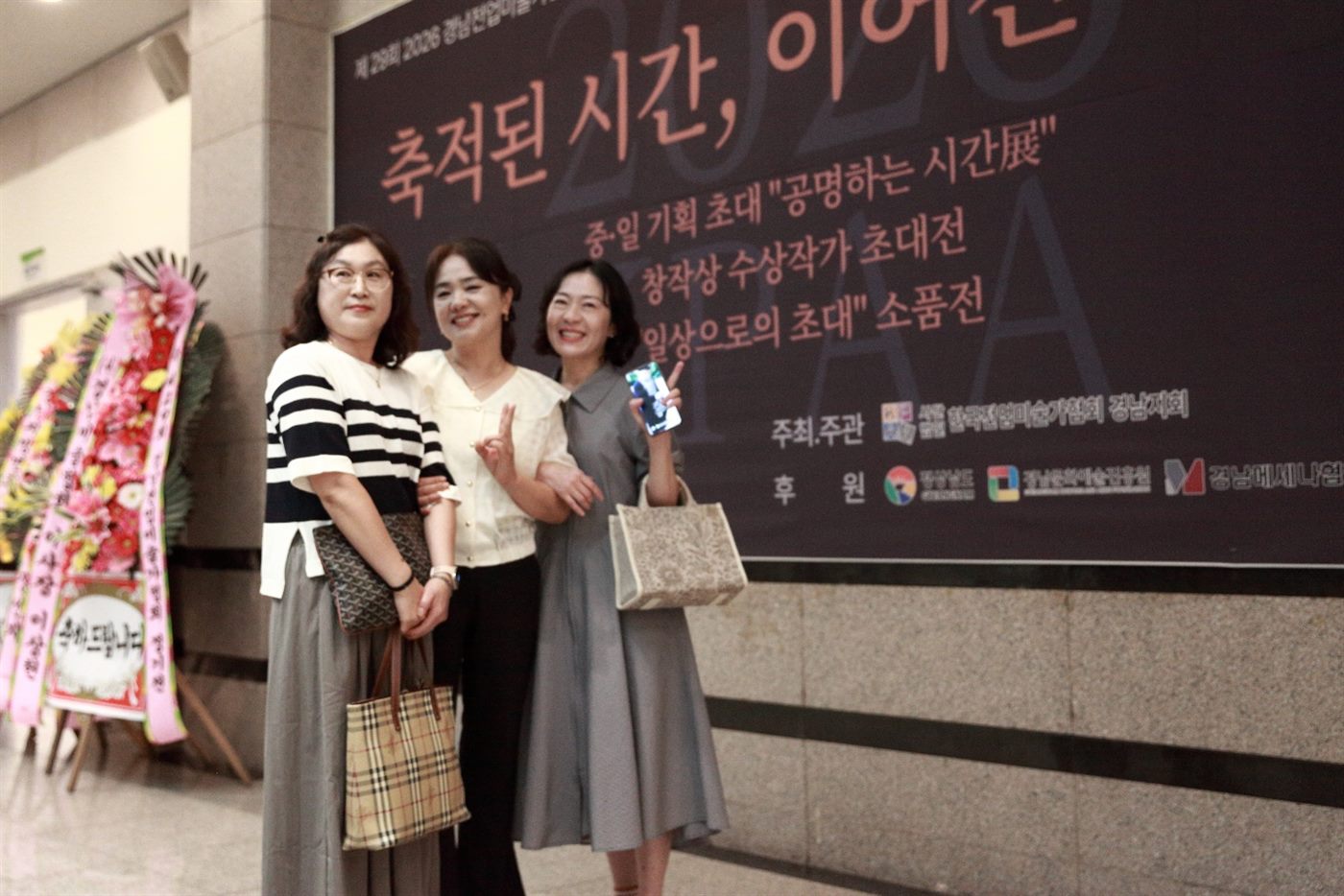 창원 성산아트홀 로비의 《축적된 시간, 이어진 시선》 전시 배너 앞에 선 세 사람