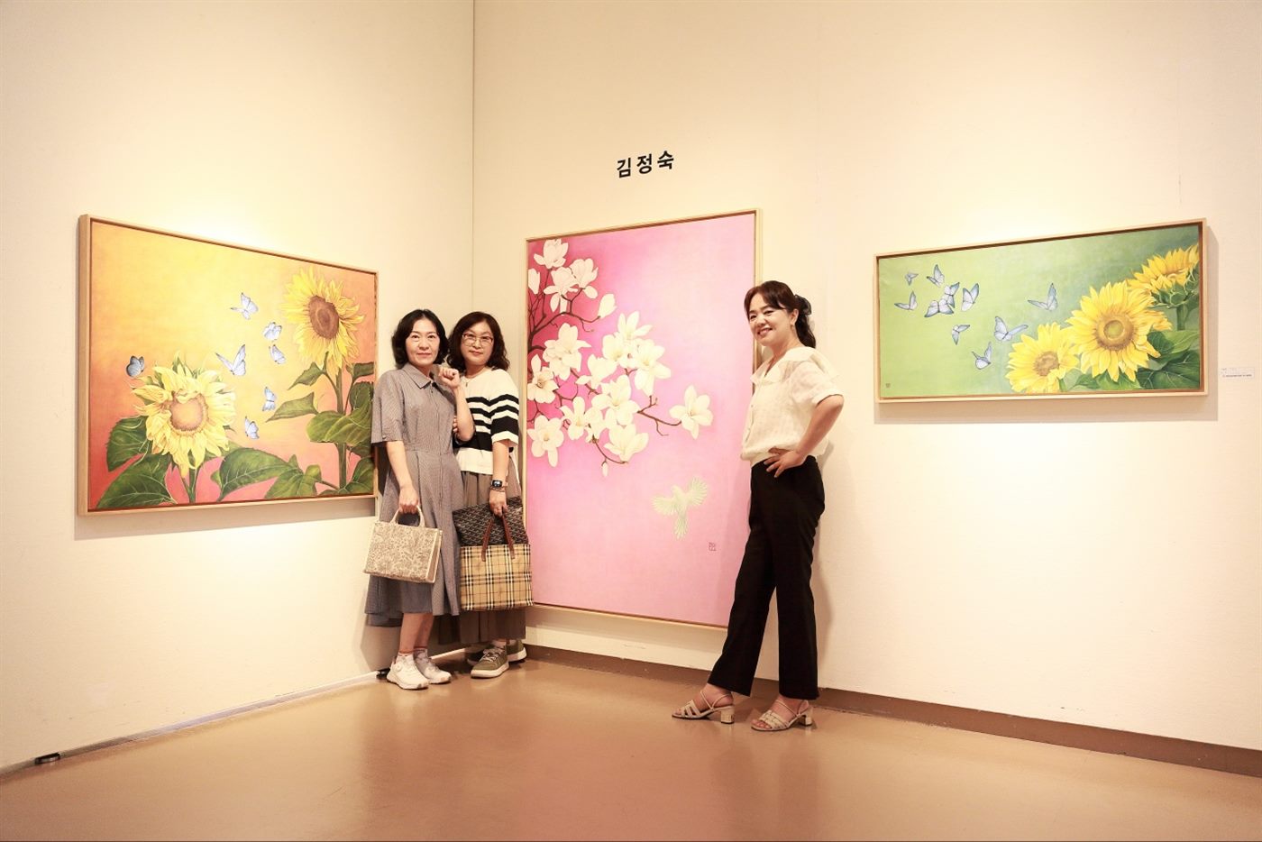 창원 성산아트홀에 마련된 김정숙 작가의 부스 앞에 선 세 사람 — 왼쪽 해바라기, 가운데 목련, 오른쪽 해바라기 작품이 걸려 있다